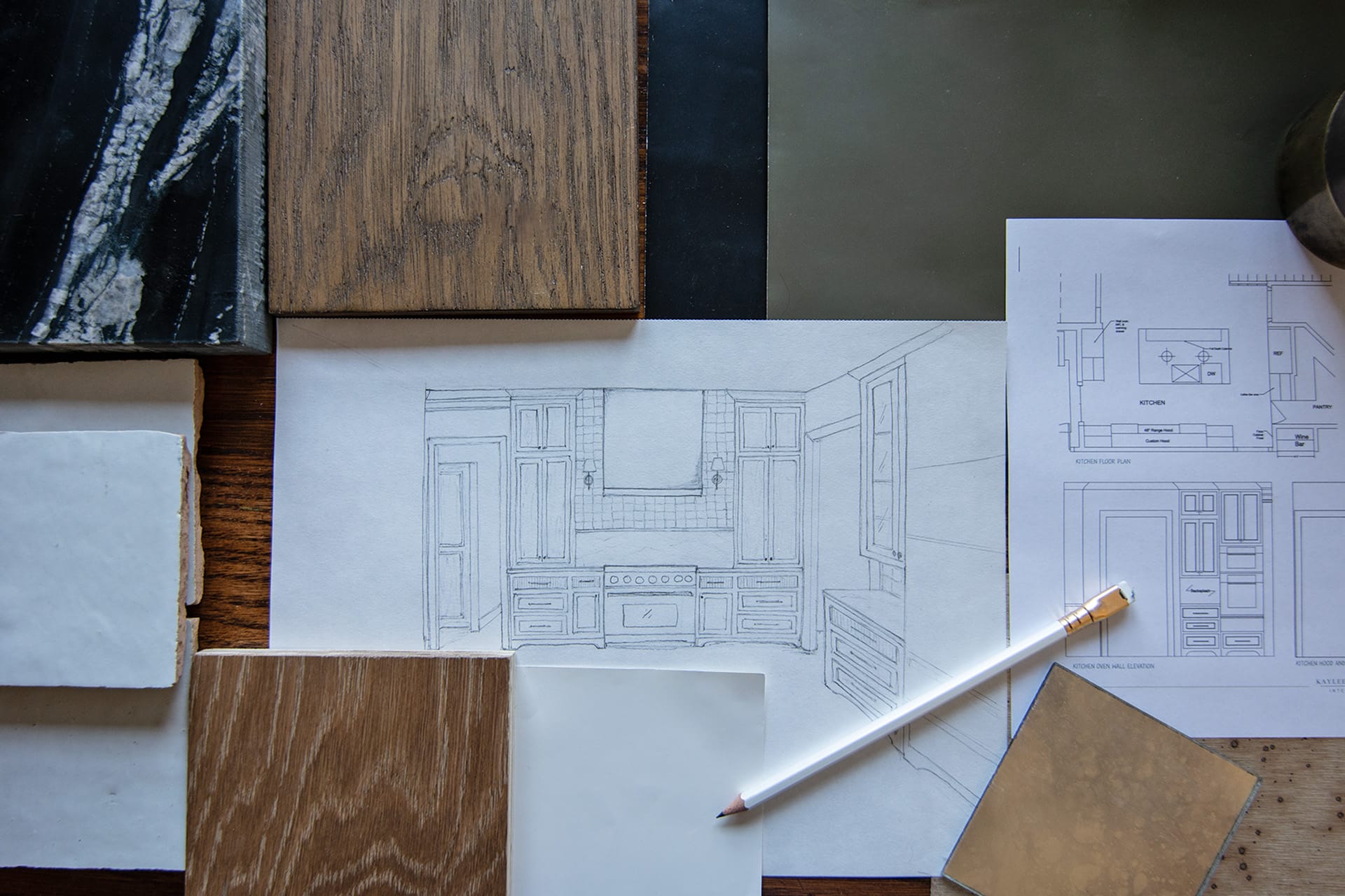 Flat lay of custom interior design solutions, including plans and material samples, featuring a kitchen sketch, wood finishes, tiles, and a pencil.