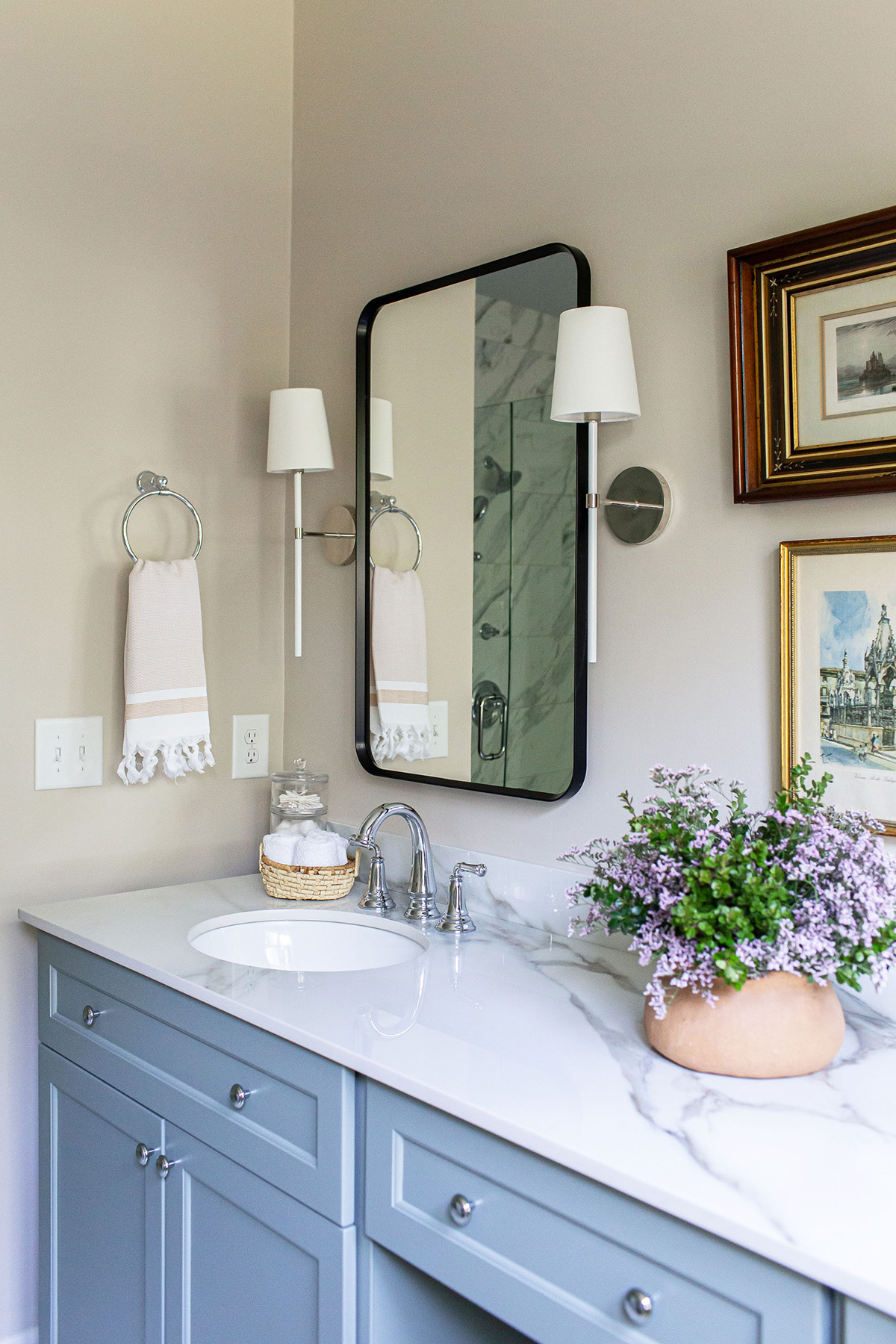 Close-up of a primary bathroom vanity with blue cabinetry, porcelain countertop, framed mirror, sconces, and floral decor.