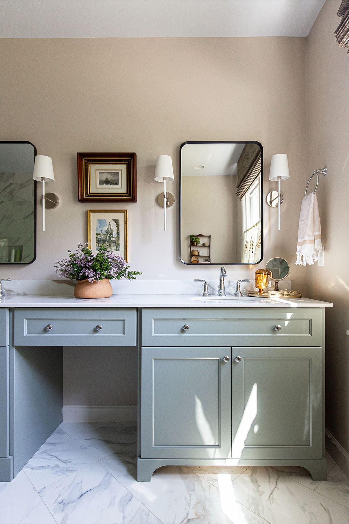 Primary bathroom vanity with blue cabinetry, porcelain countertops, framed artwork, and natural light highlighting elegant details.