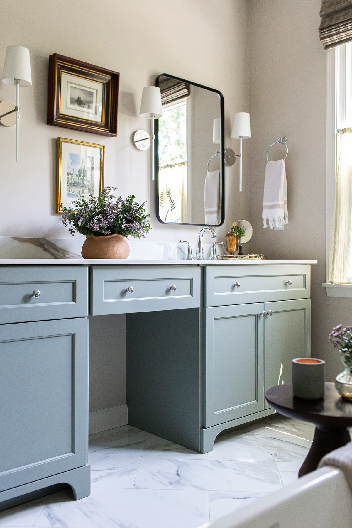 Perspective view of a primary bathroom with soft blue cabinetry, porcelain countertops, framed artwork, and elegant sconces.