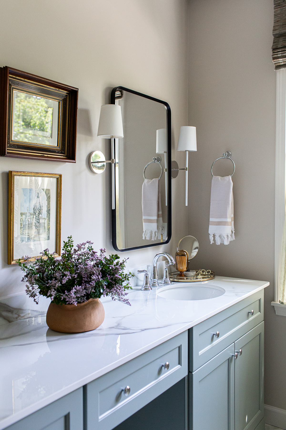 Close-up of a primary bathroom vanity with blue cabinetry, porcelain countertop, framed artwork, floral arrangement, and elegant sconces.