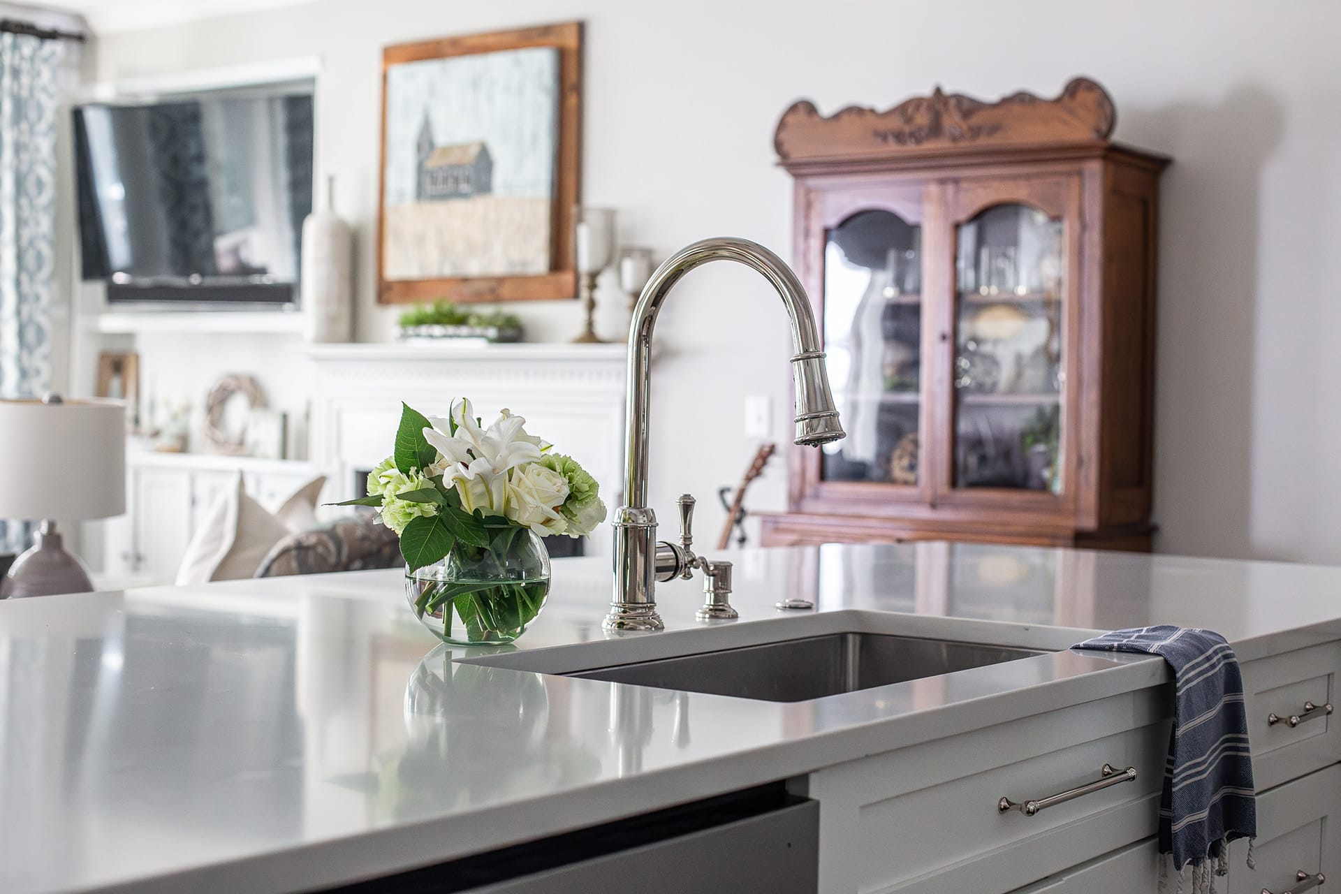 A close-up view of a white quartz kitchen island featuring a stainless steel faucet, a vase of fresh white flowers, and a cozy living space with a fireplace and vintage cabinet in the background.