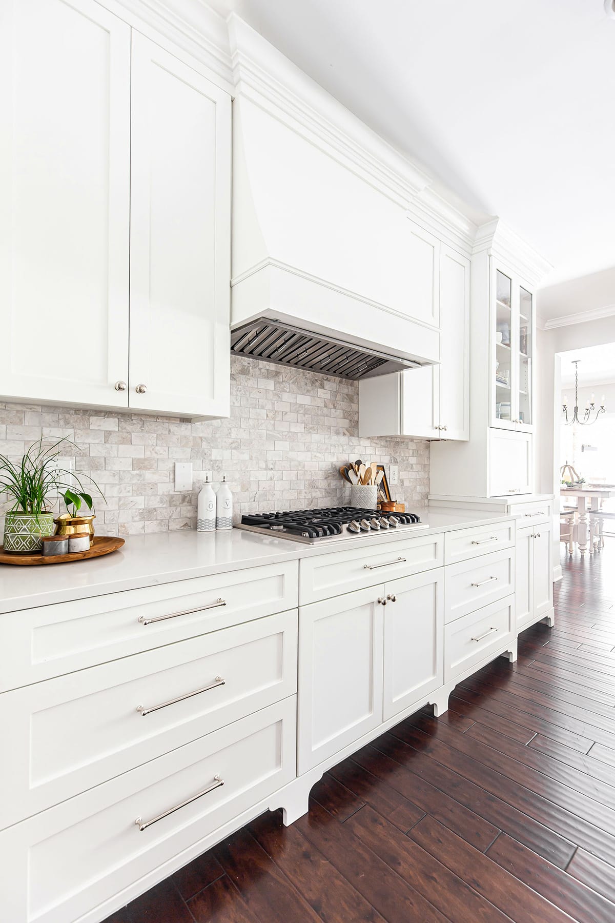 A bright kitchen with white cabinetry, a custom range hood, marble-style backsplash, and stainless steel gas cooktop, complemented by warm wood flooring.