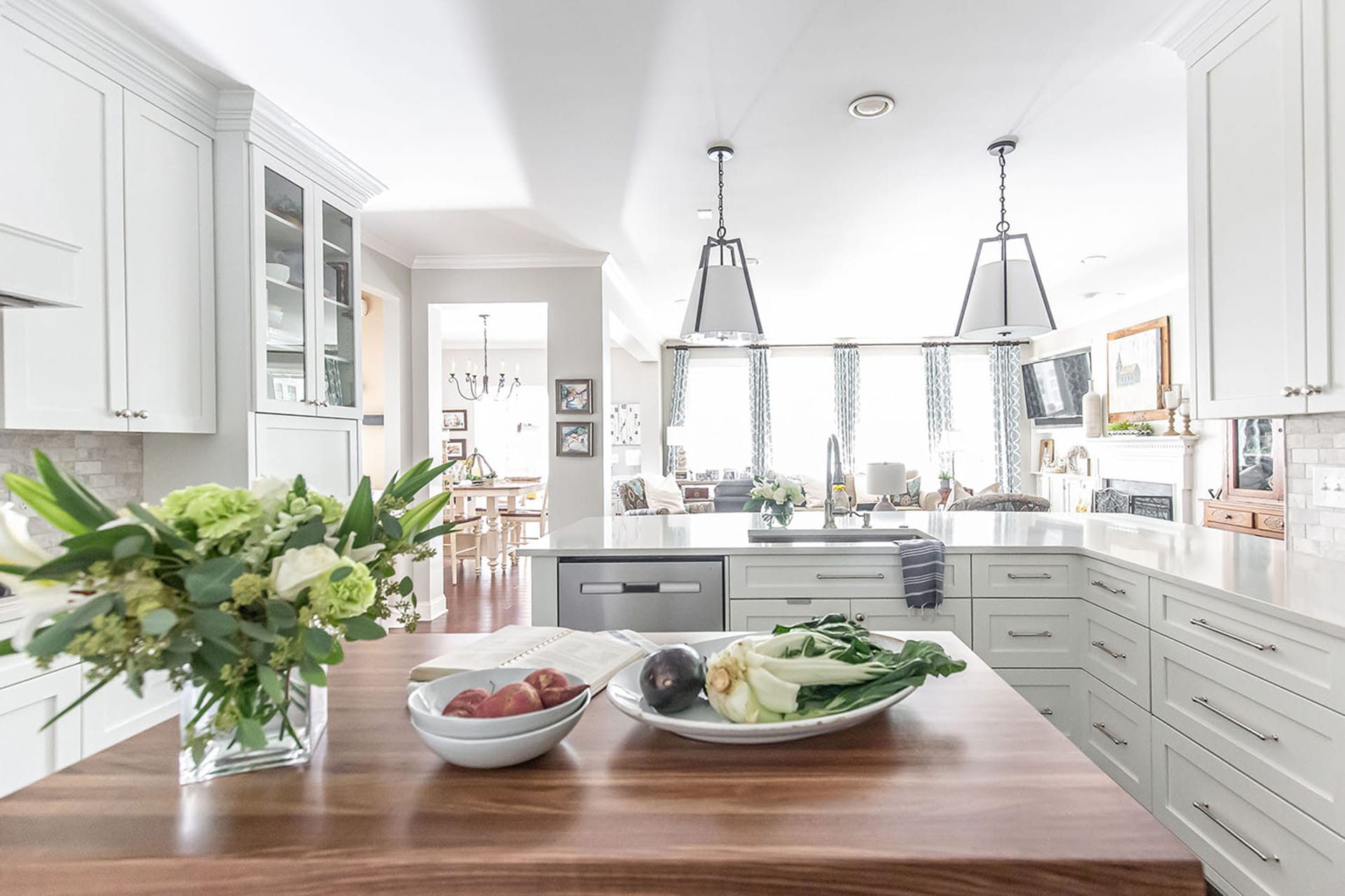 A bright and spacious kitchen tailored for modern living with white cabinetry, a large wood-topped island featuring fresh produce, pendant lighting, and an open view into a cozy living and dining area.