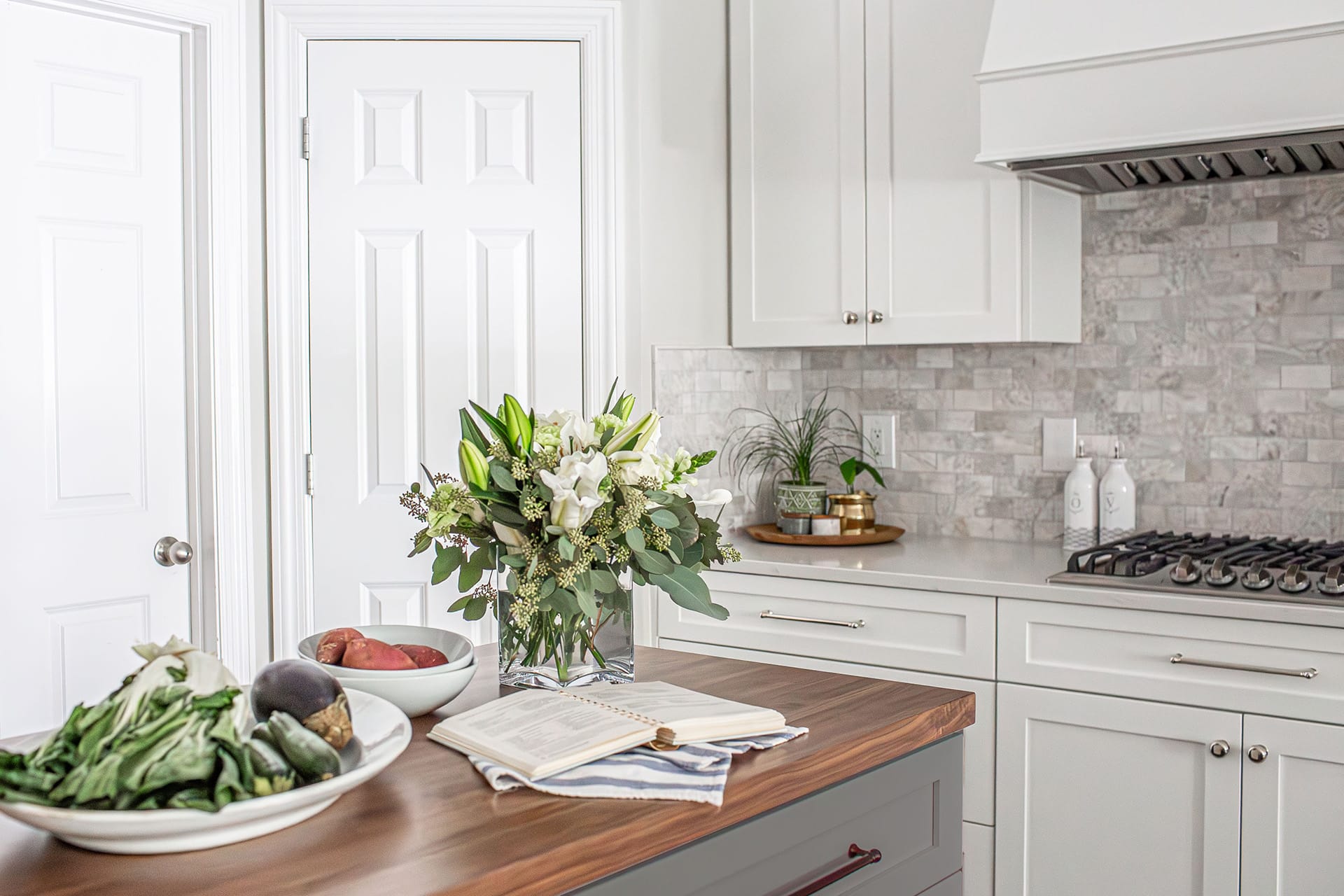 A bright kitchen with white cabinetry, a wood-topped island featuring a bouquet of flowers, fresh produce, and an open cookbook, set against a marble-style backsplash and a gas cooktop.