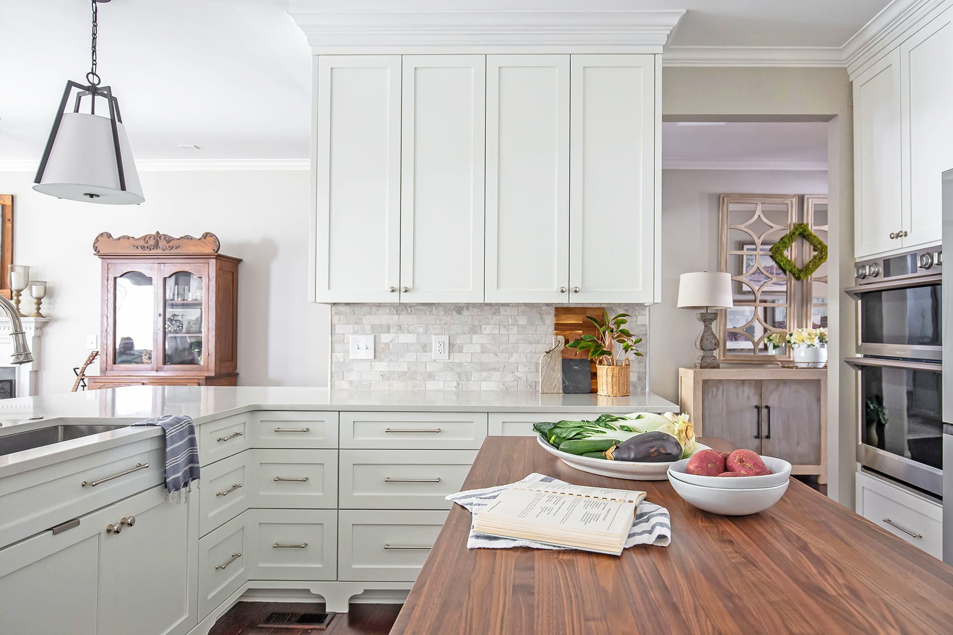 A kitchen featuring white cabinetry, a warm wood-topped island styled with fresh vegetables and an open cookbook, set against a marble-style backsplash with adjacent living space details.