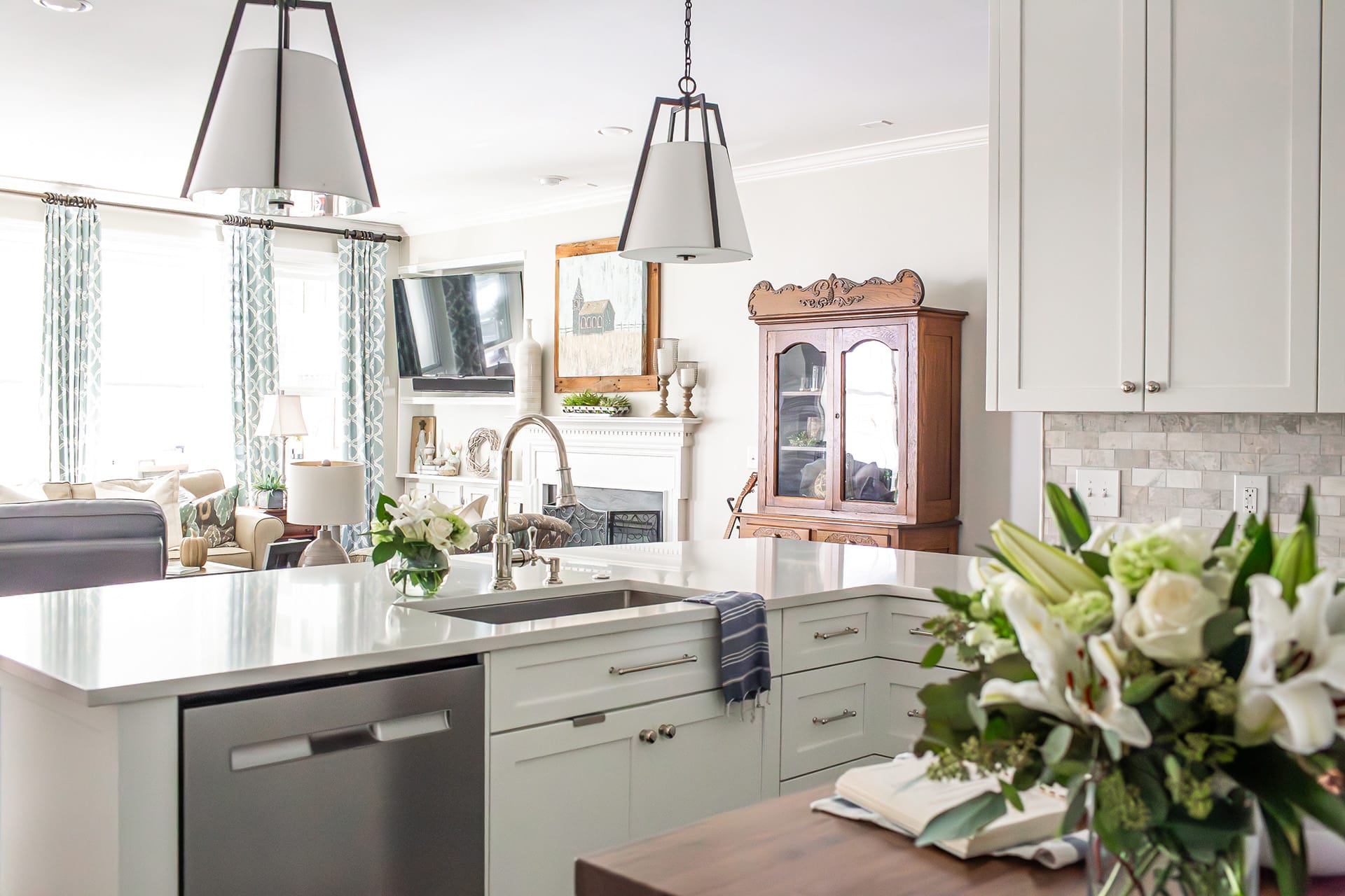 A bright kitchen with a white quartz island featuring a stainless steel sink, pendant lighting, and a view into a cozy living room with traditional decor elements.