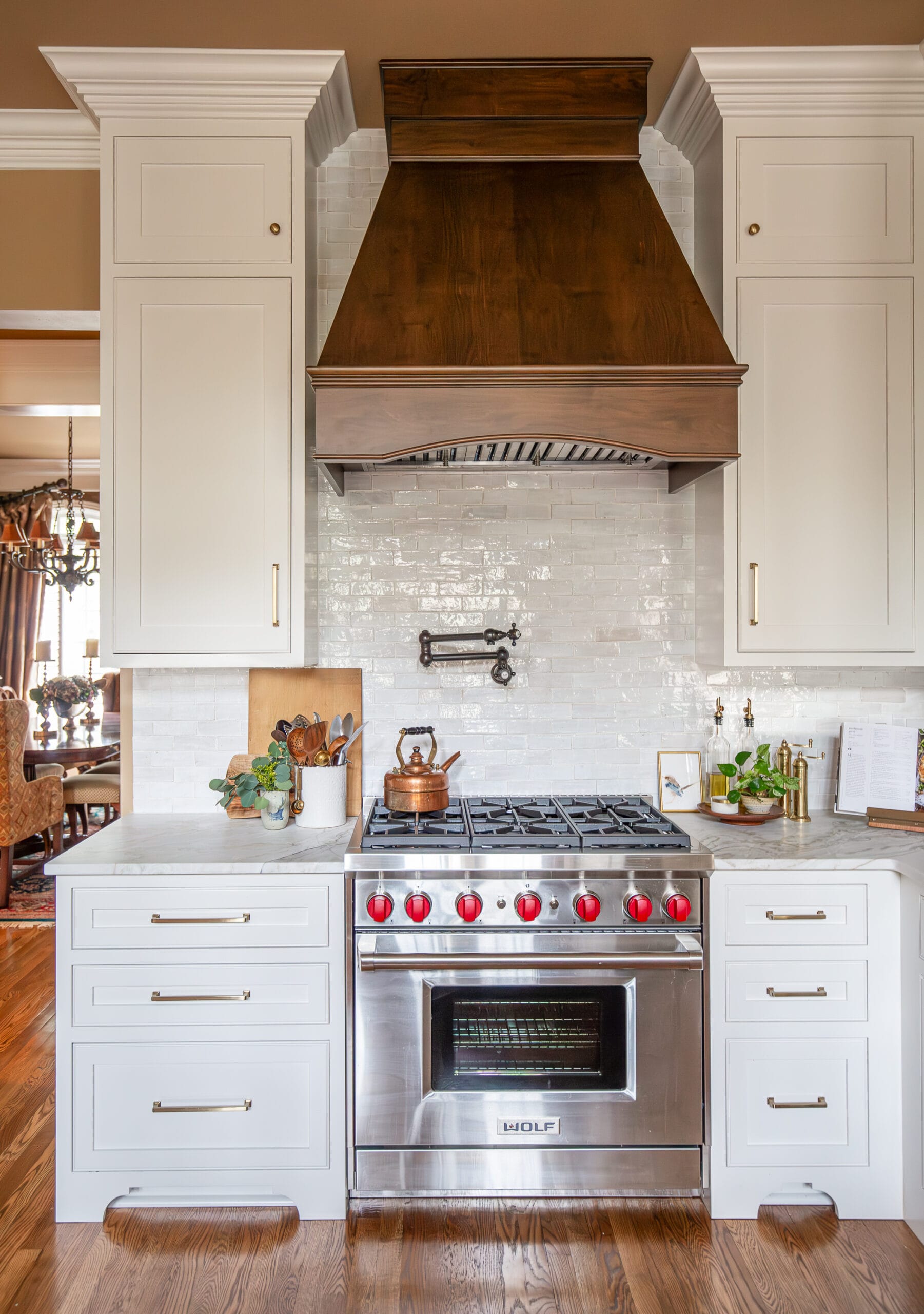 Elegant kitchen with custom wooden hood, white cabinetry, marble countertops, and a stainless steel Wolf range with red knobs.